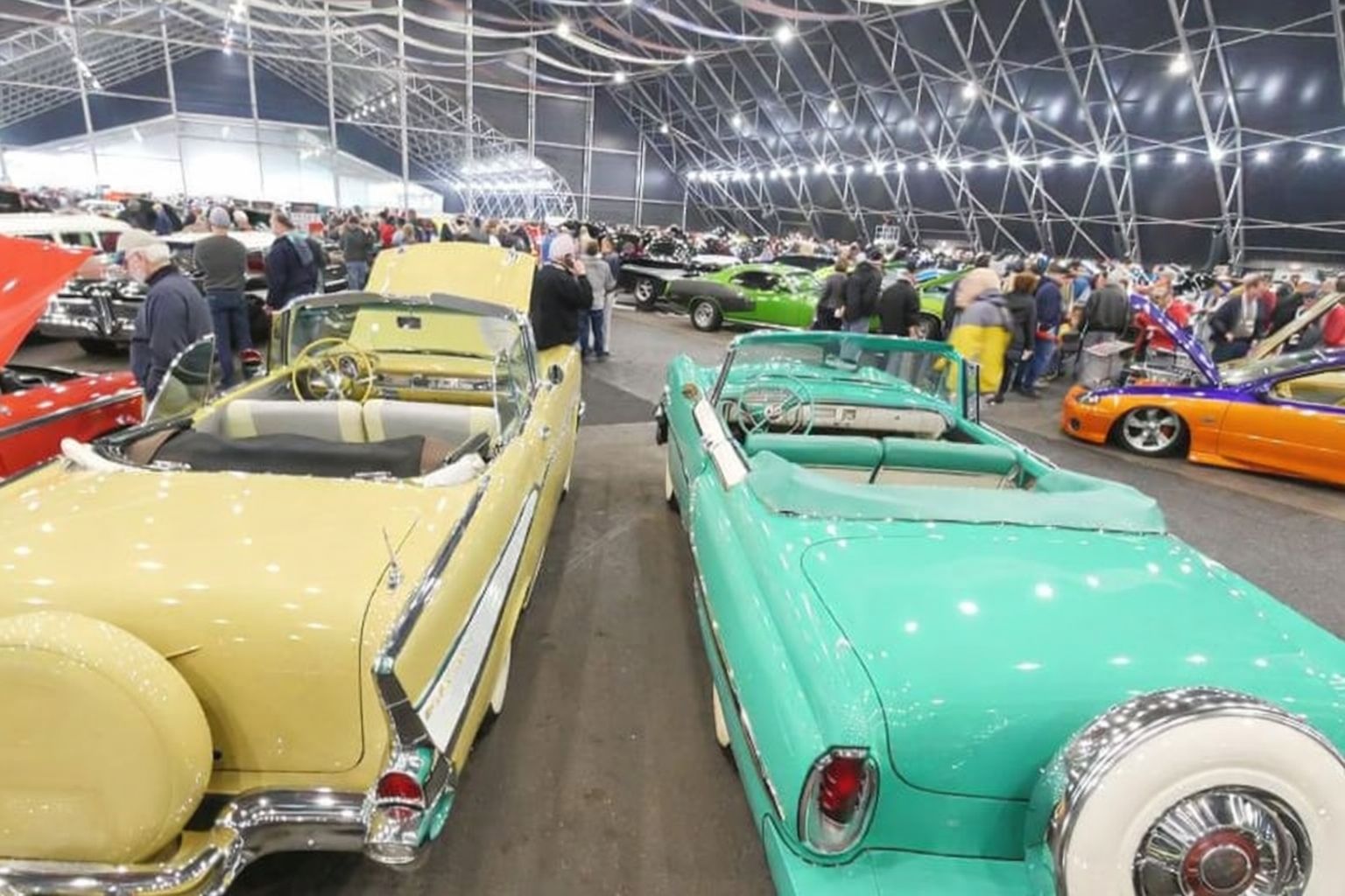 Två veteranbilar, gul och turkos, står på en fordonsauktion med många besökare.